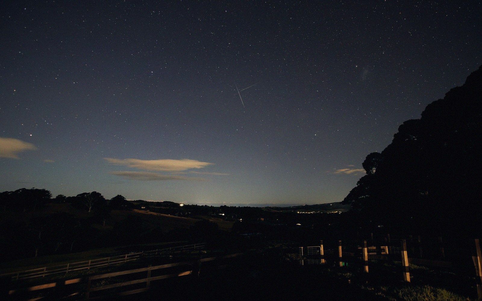 A faint meteor streak crosses a starry night sky above dark rolling hills and a fence